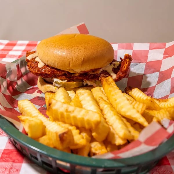 Spicy fried chicken sandwich with pickles and sauce, served with crinkle-cut fries in a basket.
