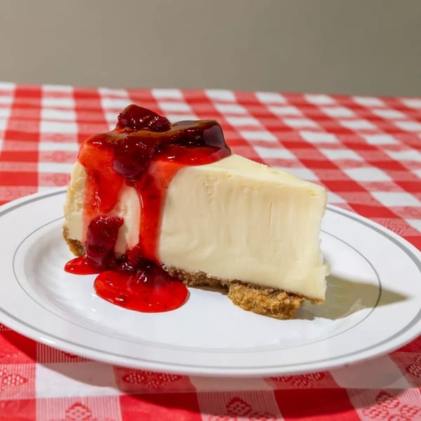 Slice of cheesecake with strawberry topping on a white plate, on a red and white checkered tablecloth.