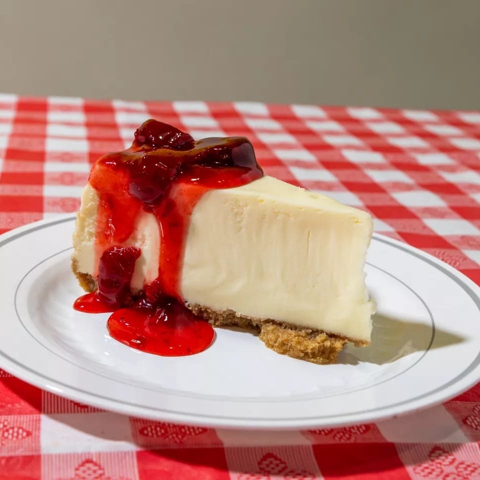 Slice of cheesecake with strawberry topping on a white plate, on a red and white checkered tablecloth.