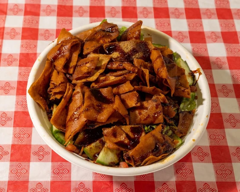 Fattoush salad with crispy pita bread and dark dressing in a white bowl on a red and white checkered tablecloth.