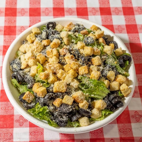 A white bowl of green salad with black olives, croutons, and grated cheese on a red and white checkered tablecloth.
