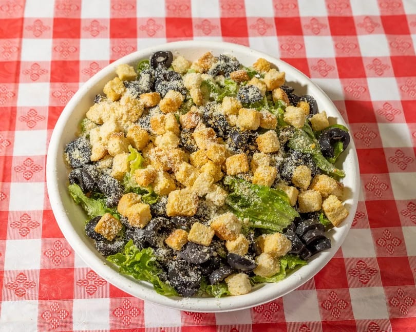 A white bowl of green salad with black olives, croutons, and grated cheese on a red and white checkered tablecloth.